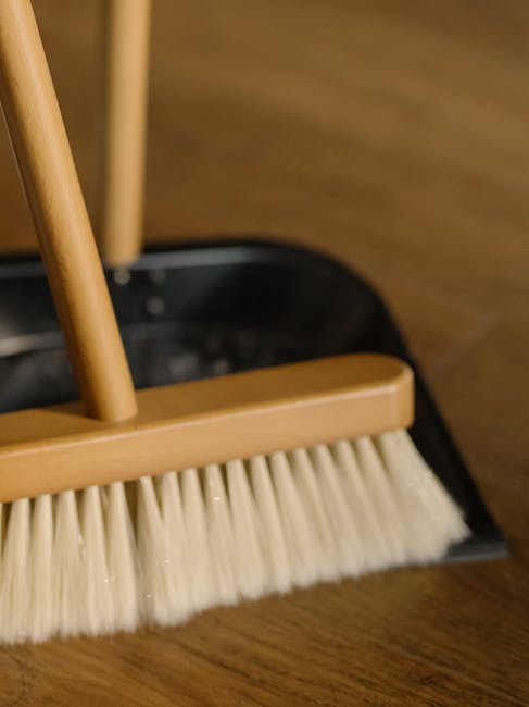 Close-up image of a broom with a wooden handle and cream-colored bristles, positioned on a polished wooden floor within a room. The broom appears to be part of a cleaning process, with a dustpan visib