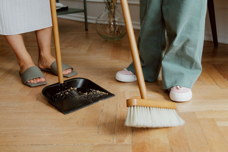 Close-up image of a broom with a wooden handle and cream-colored bristles, positioned on a polished wooden floor within a room. The broom appears to be part of a cleaning process, with a dustpan visib